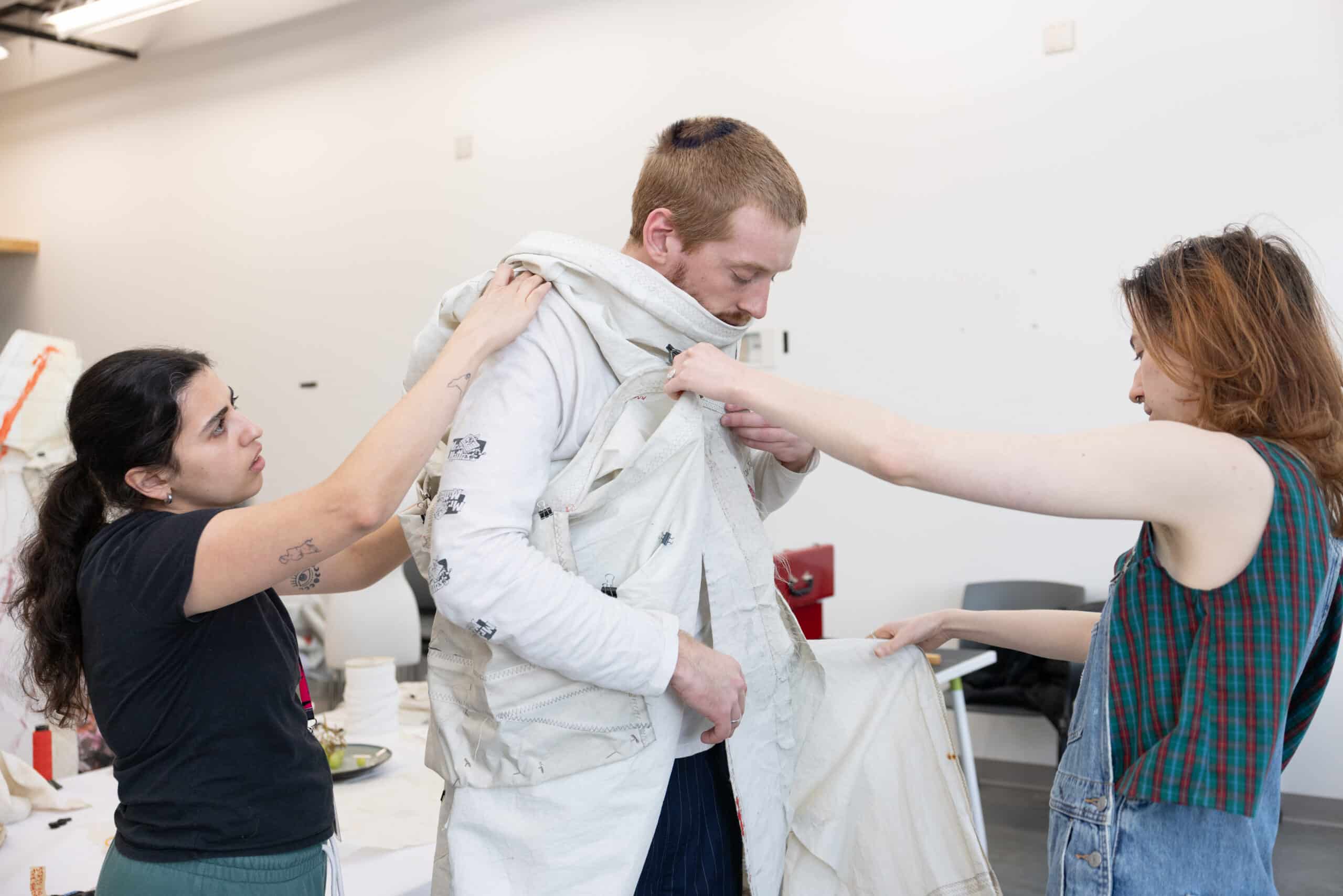 Two people help fit a layered garment on another, hands steady at seams and closures, shaping fabric into form.