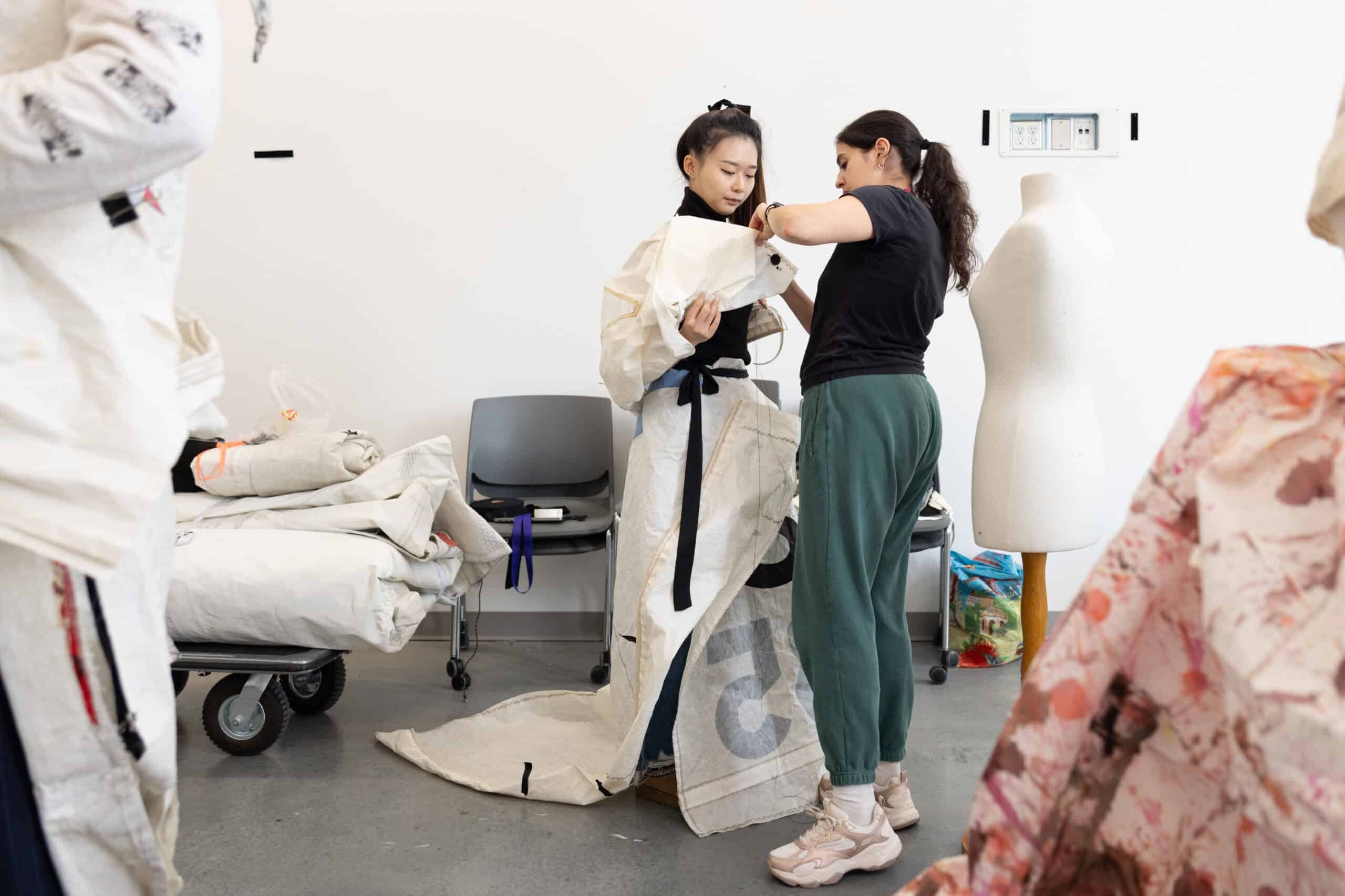 In a busy studio, one person is fitted while others move between tasks, fabric draped across tables and forms like unfinished ideas.