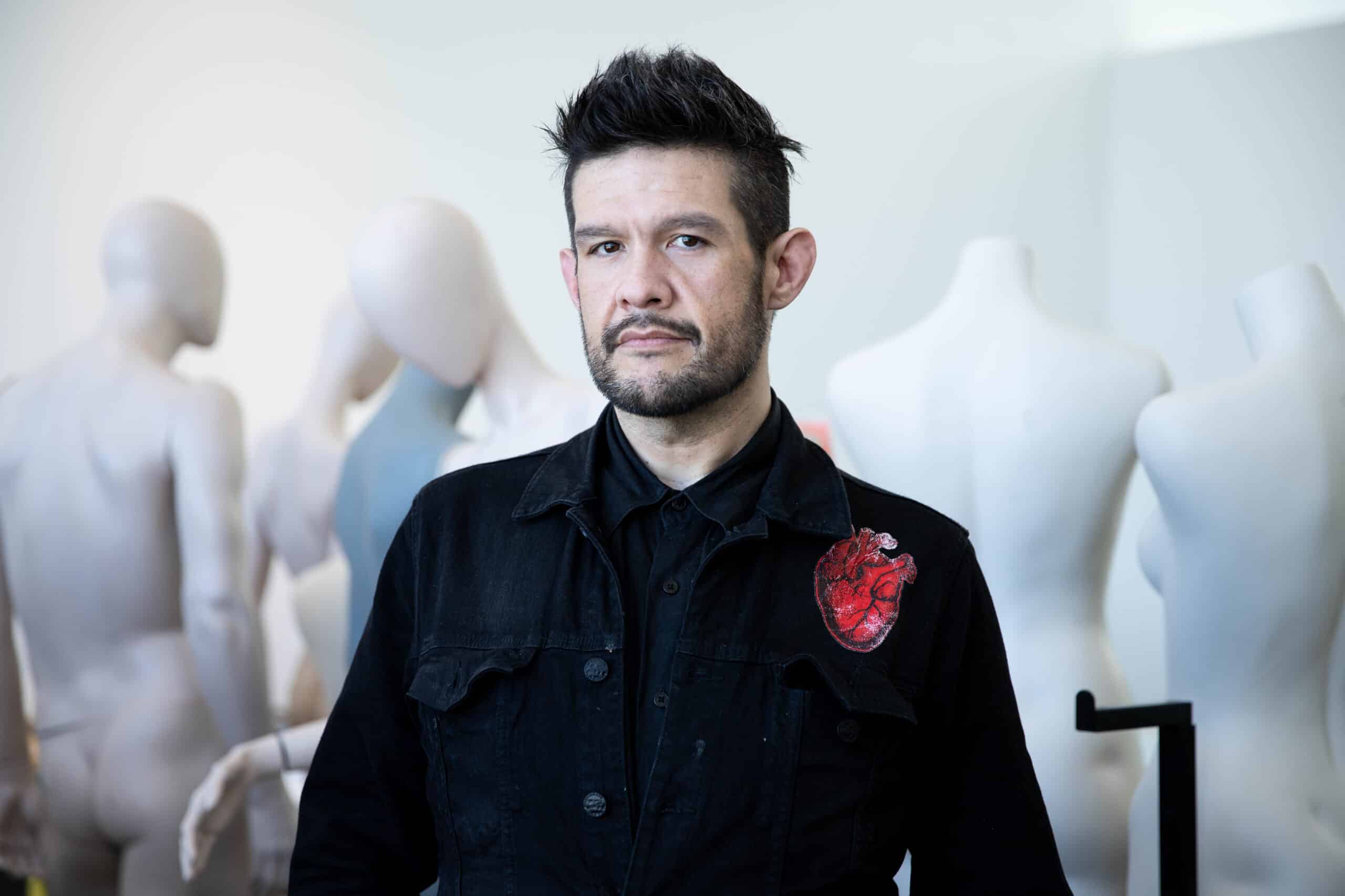 A person stands before a row of blank dress forms, wearing a dark jacket marked with a red heart; their steady gaze meets the camera, the mannequins behind them like silent witnesses.