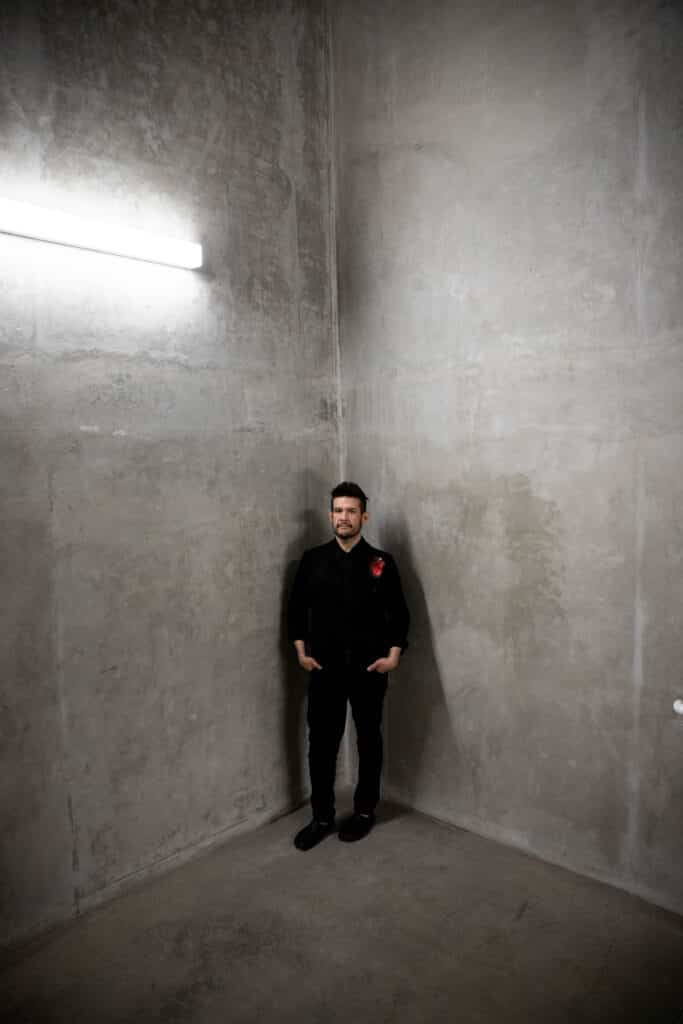 In a tall, concrete corner, a lone figure stands with hands in pockets beneath a bright wall light, their presence small against the textured gray expanse.