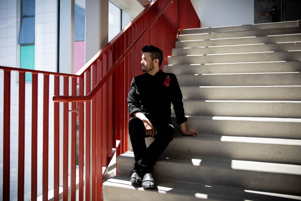 Seated on wide concrete steps beside a bold red railing, a person looks off into the light, their dark clothing catching the sharp contrast of sun and shadow.