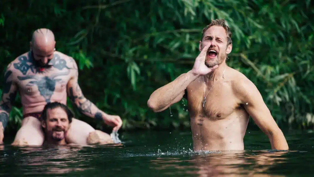 In shallow water, one person cups their hands to call out while others gather nearby, bodies half-submerged, sharing a lively, fleeting moment against a backdrop of green