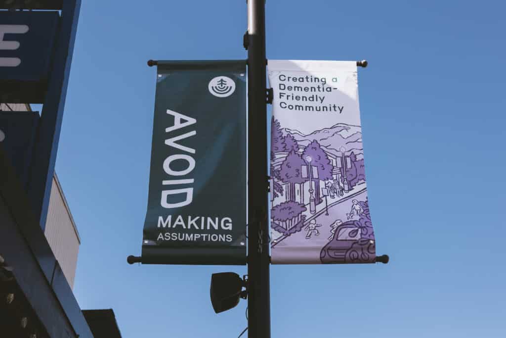 Two street banners hang from a pole against a clear blue sky—one reading “Avoid Making Assumptions,” the other “Creating a Dementia-Friendly Community,” illustrated with a gentle neighbourhood scene.