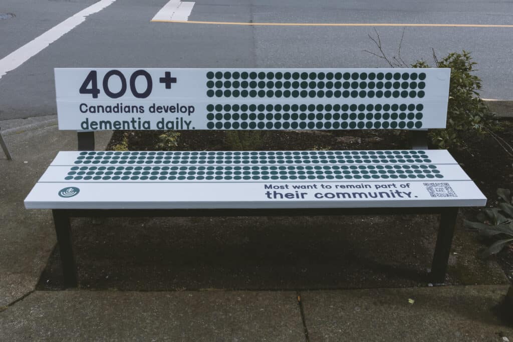 A public bench printed with rows of green dots and the message “400+ Canadians develop dementia daily” sits on a sidewalk, inviting pause and reflection on community and care.