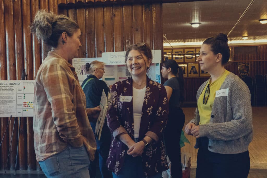Three people stand in conversation inside a wood-paneled hall, name tags visible, their expressions open and engaged as posters about dementia-friendly environments line the background.