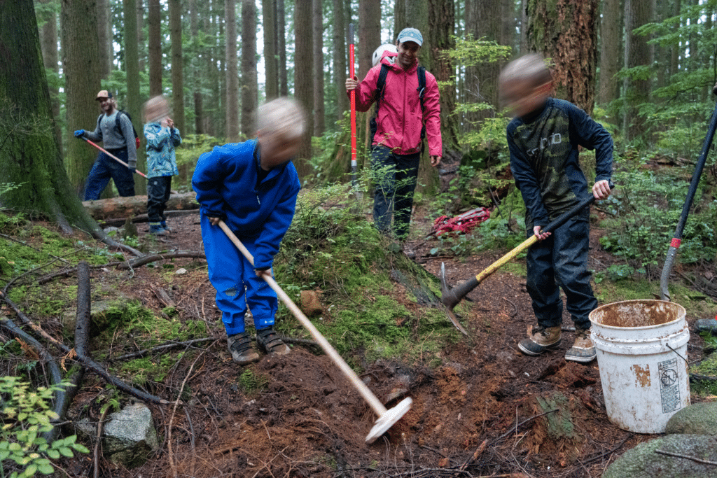 Children and adults work together along a forest trail, digging into damp earth beneath tall cedar trees as tools and conversation move through the rain-soaked woods.