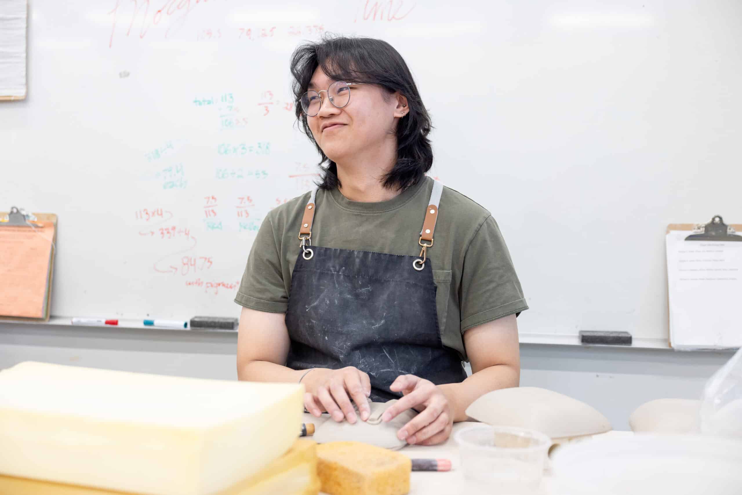 A ceramics student sits at a studio table with clay-covered hands, glancing upward as handwritten notes and measurements cover the whiteboard behind them.