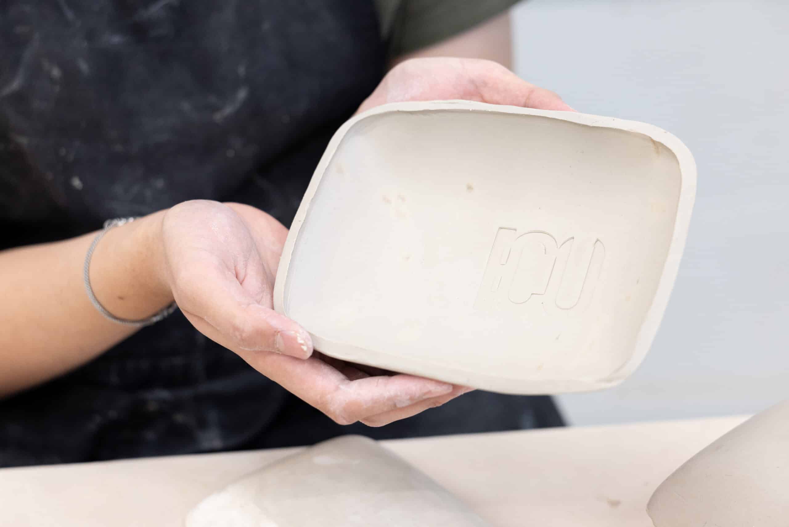 A student holds a freshly formed ceramic dish toward the camera, its smooth interior marked with raised lettering pressed into the clay.