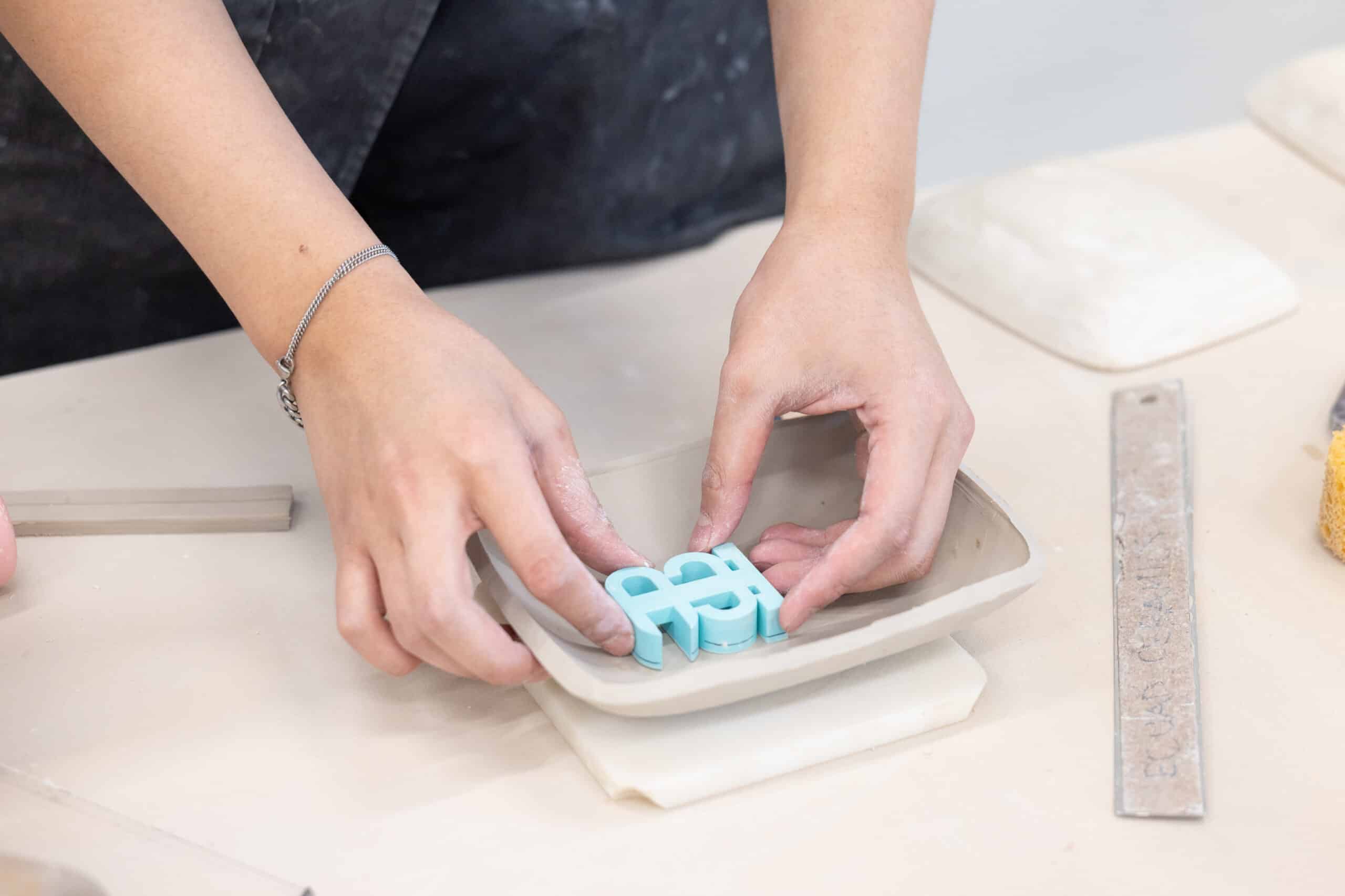 Close-up of clay-dusted hands carefully placing blue letterforms into a shallow ceramic dish on a studio table.