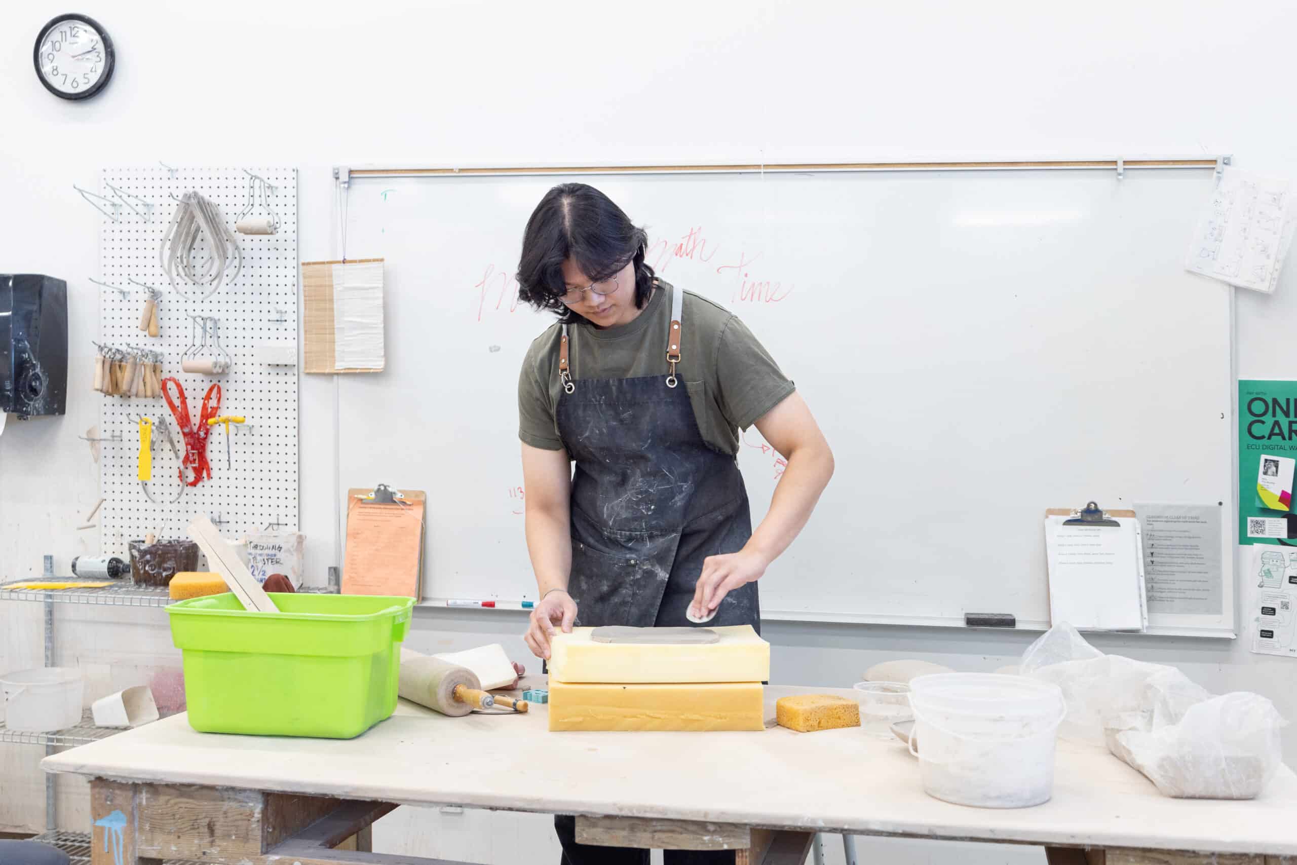 A ceramics student shapes a shallow clay form on layered foam supports in a bright studio lined with shelves, tools and handwritten notes.