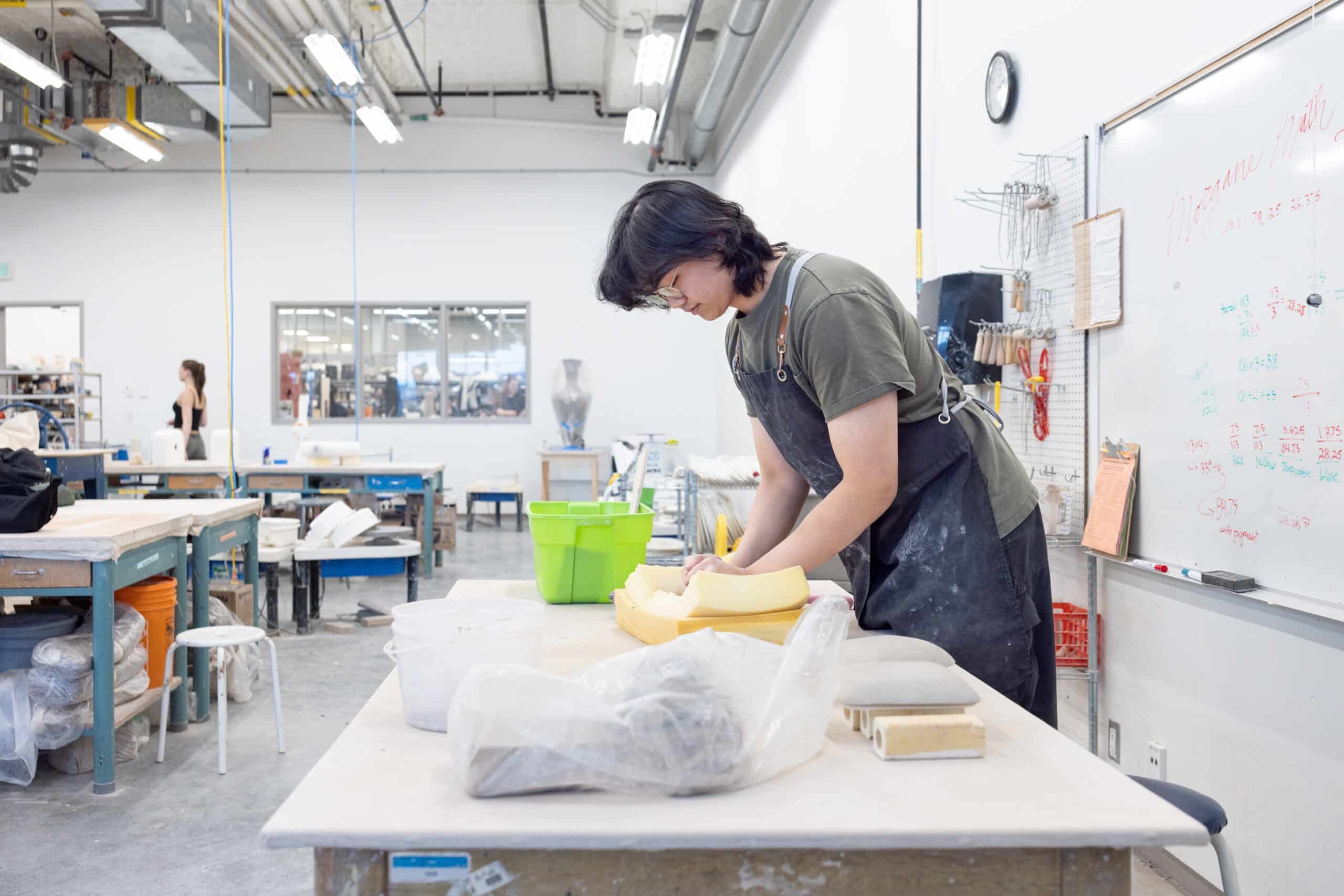 A ceramics student bends over a long studio table, shaping clay by hand beneath bright overhead lights in a spacious workshop.