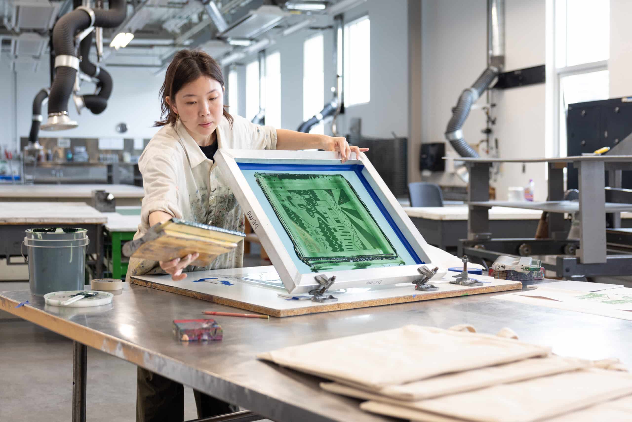 A student lifts a screen-printing frame coated in green ink, preparing the next print on a large studio worktable scattered with tools and paper.