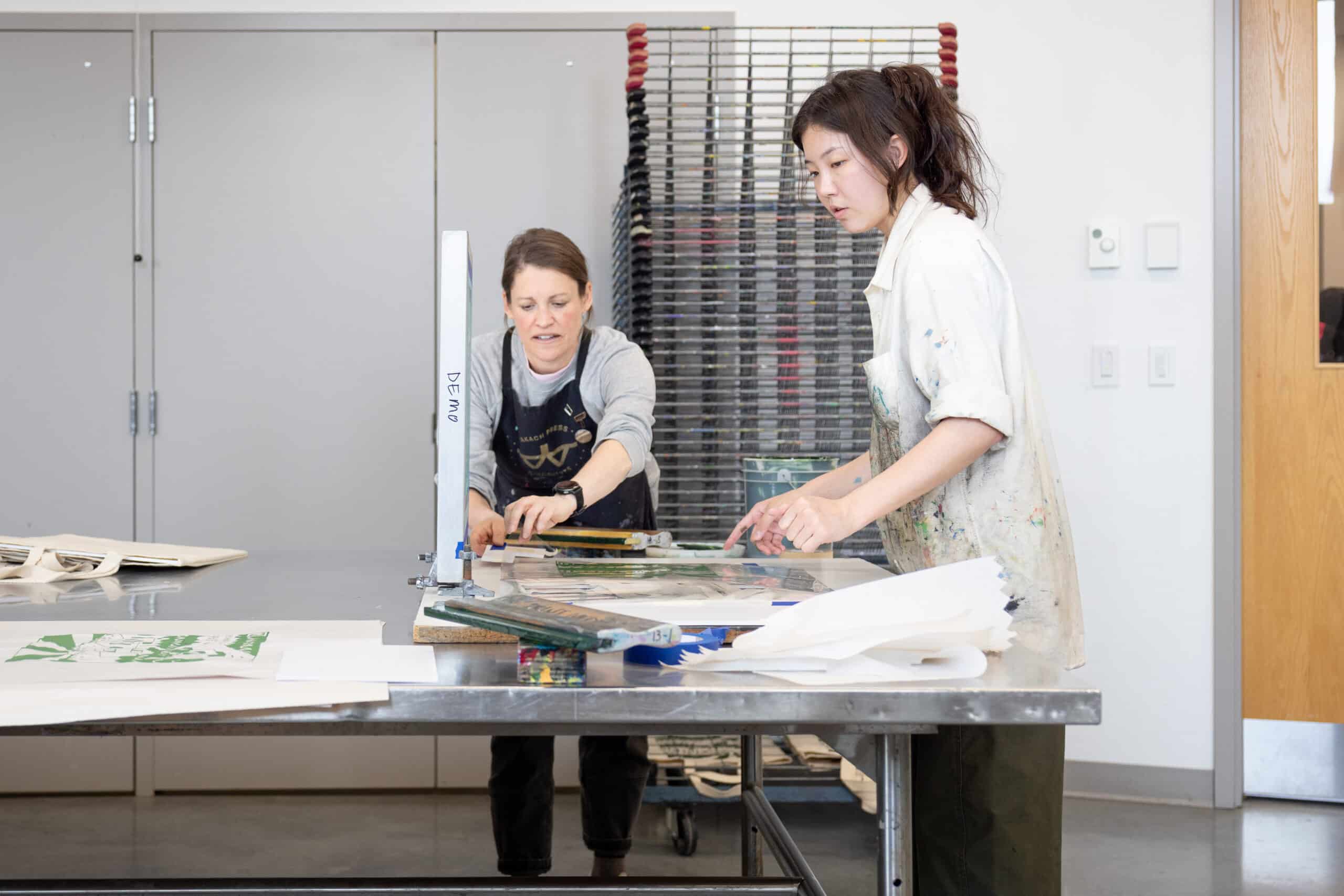 Two people lean over a screen-printing station, pointing to details on the mesh frame as paper and tools gather across the studio table around them.