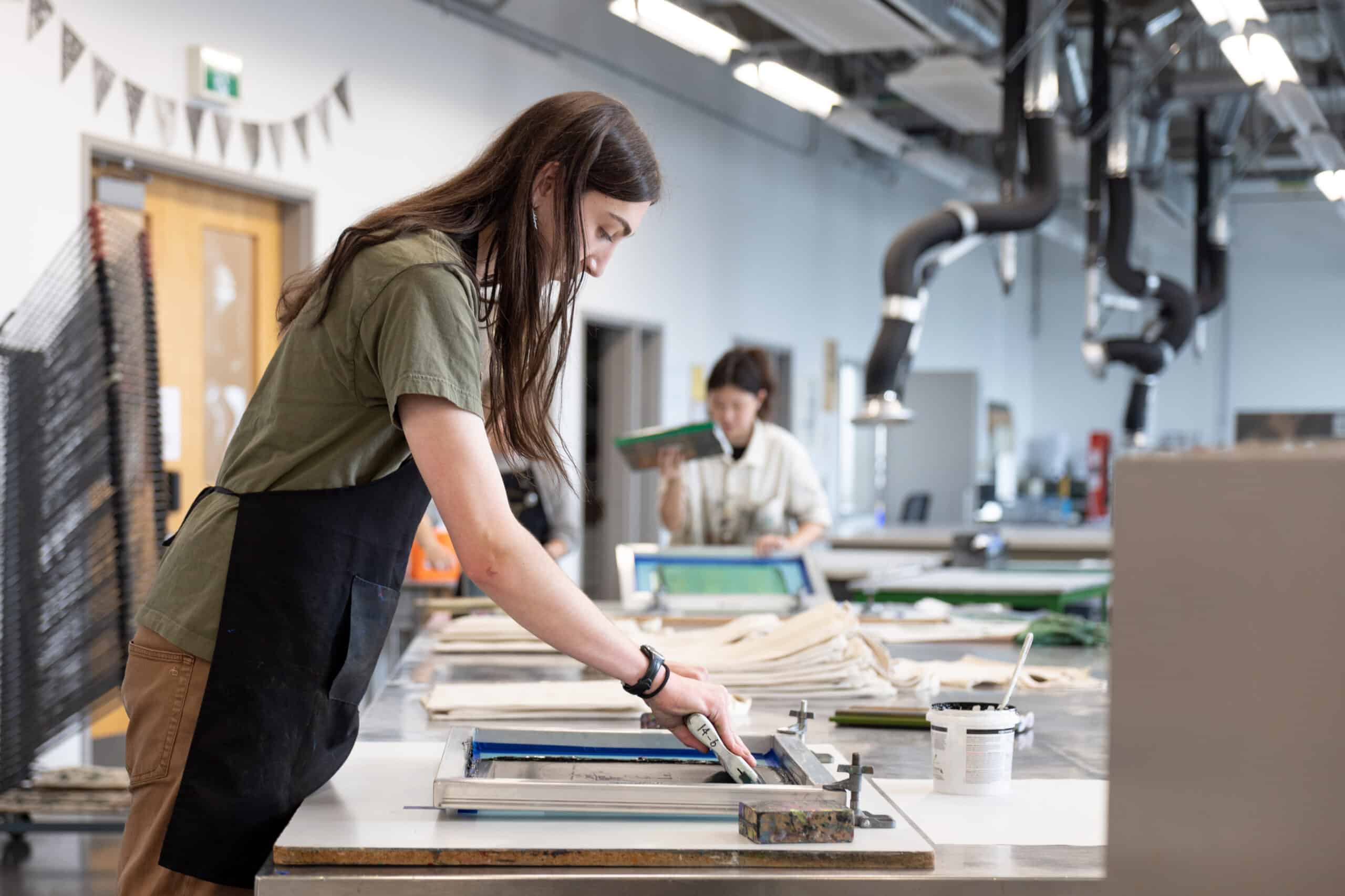 A student in a dark apron pulls a squeegee across a screen-printing frame while another student works in soft focus farther down the studio table.