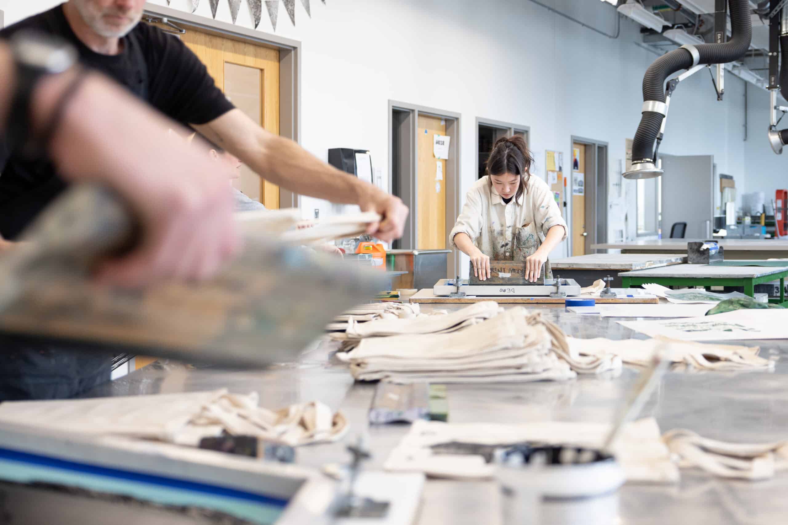 In a busy print studio, one student lifts a printing screen while another presses ink across a frame, surrounded by stacks of unfinished tote bags.