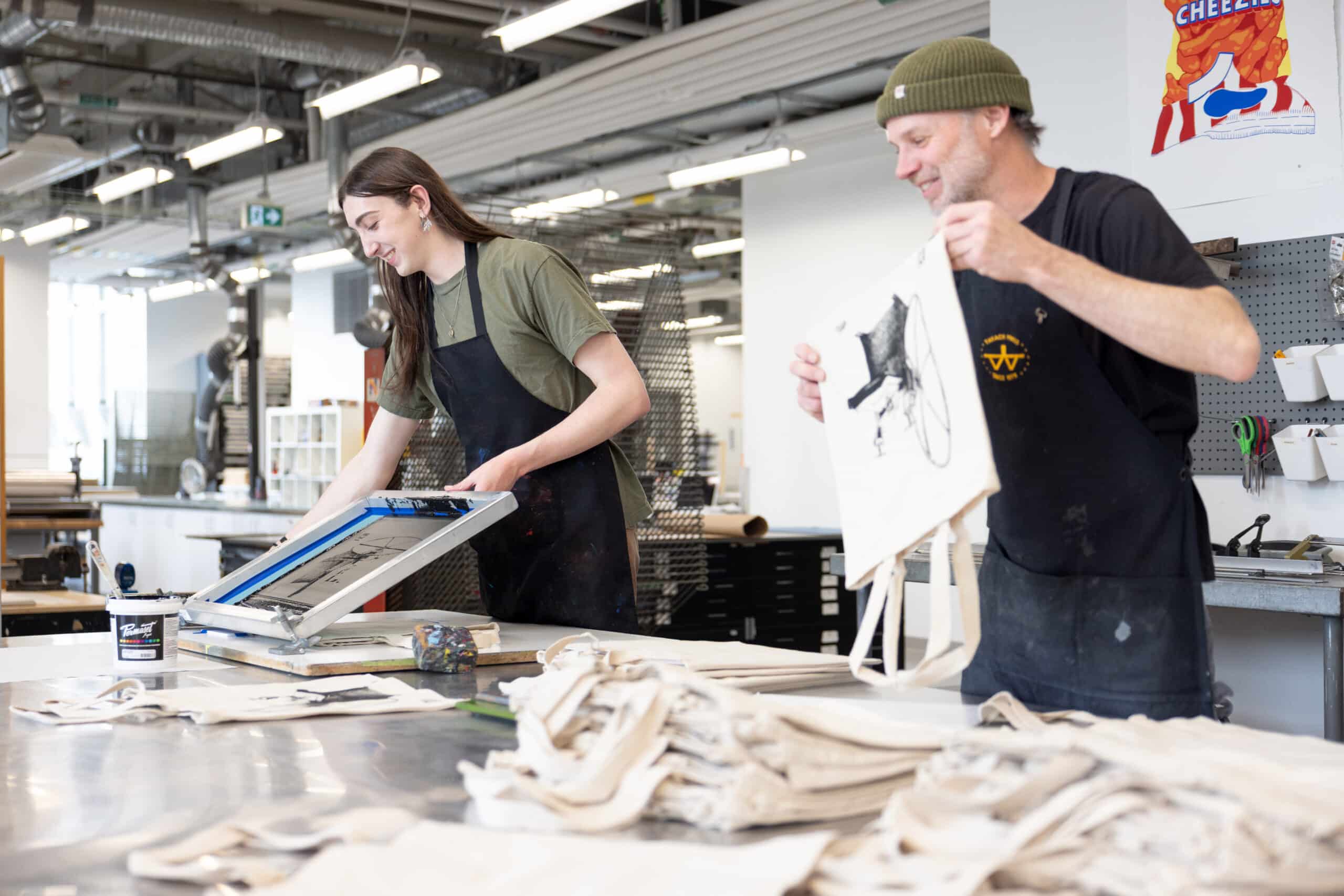 Two people smile while revealing a freshly printed tote bag in a bright studio filled with screens, ink and rows of drying racks.