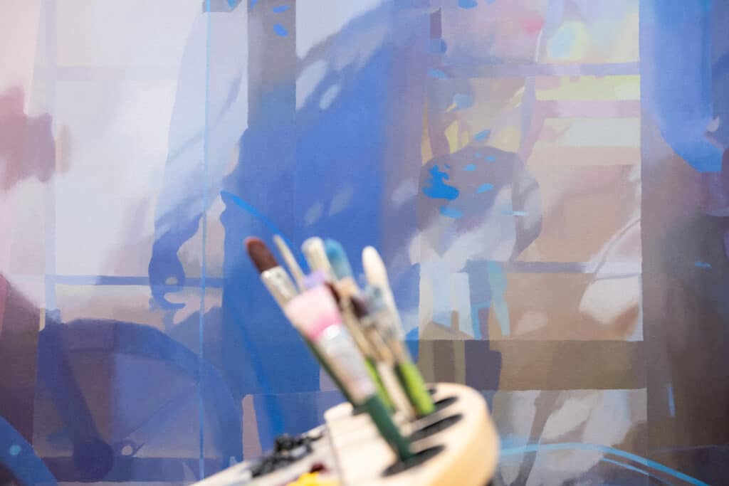 Paintbrushes rest in the foreground against a blurred abstract painting filled with deep blues, soft purples and translucent shapes.