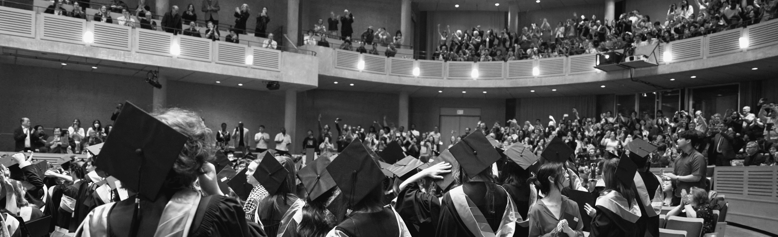 Black-and-white photograph of a convocation ceremony viewed from behind a crowd of graduating students in caps and gowns. Graduates fill the foreground while family, friends and supporters stand and applaud from curved balcony levels above, creating a celebratory atmosphere in a large auditorium.