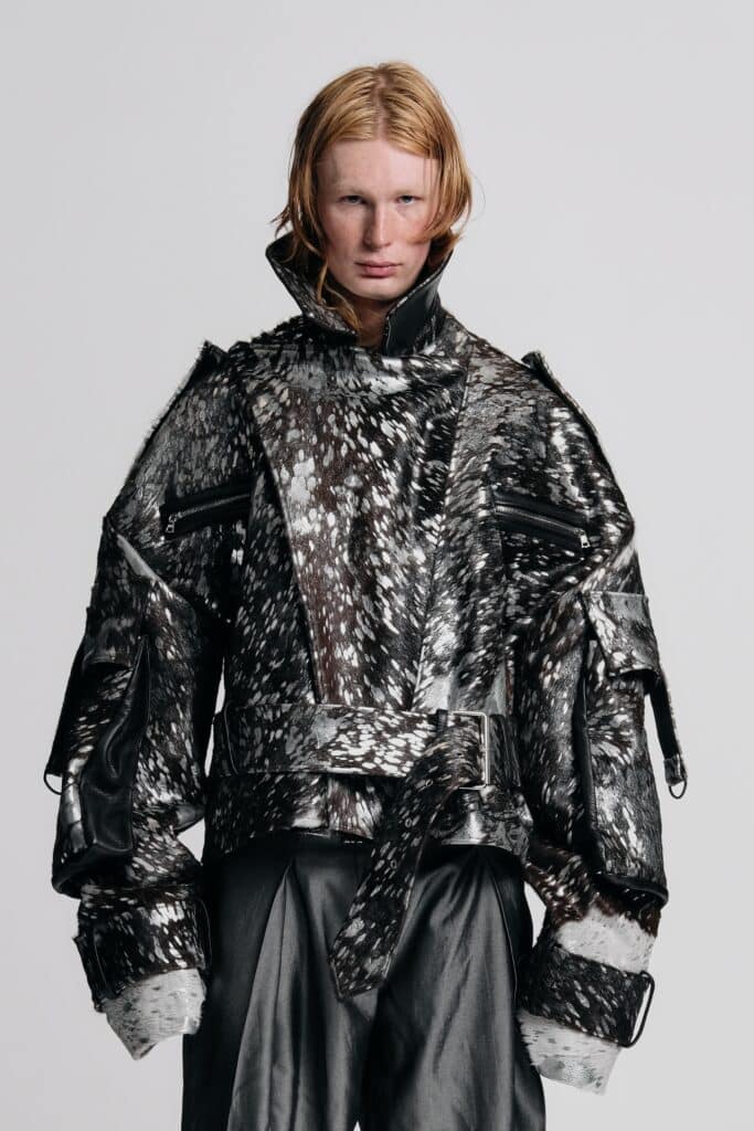 A model stands against a light grey backdrop wearing an oversized black-and-silver textured jacket with exaggerated sleeves, wide leather trousers and a belted waist, gazing directly at the camera.