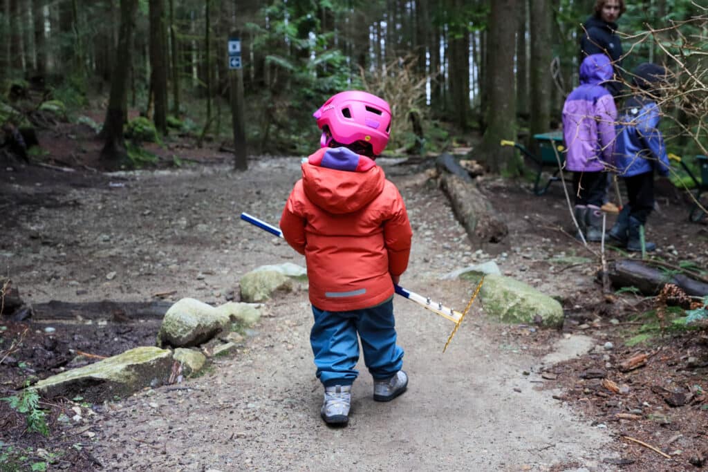 Child in a bright pink helmet walks down a wooded trail carrying a rake, moving toward others gathered beneath tall evergreens.