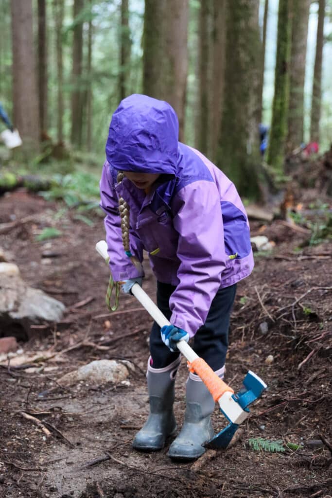 Child in a purple rain jacket grips a trail tool and shapes wet earth along a forest path, surrounded by towering trees and moss-covered ground.