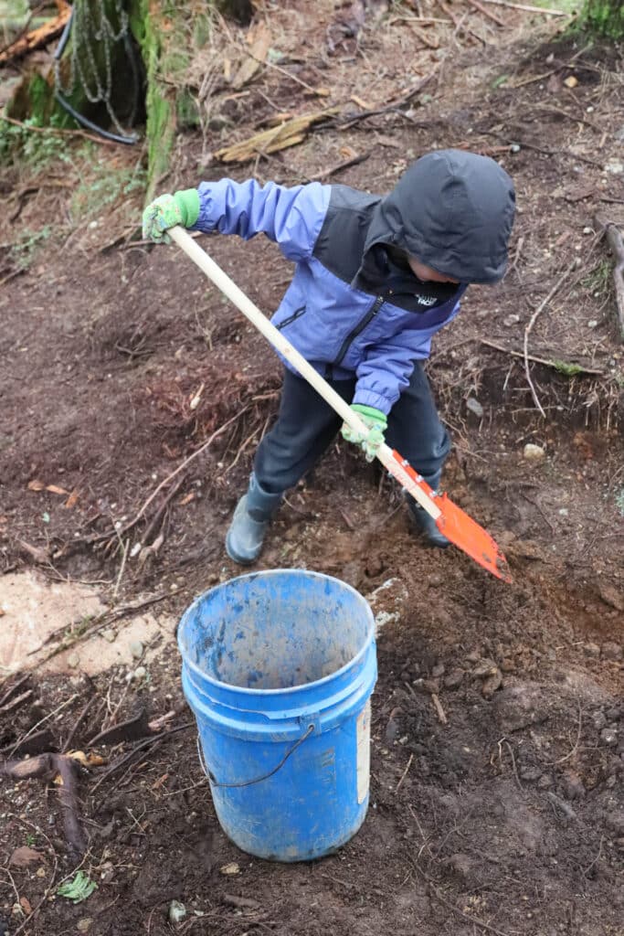 Child in rain boots digs carefully into damp soil with a small shovel beside a blue bucket, carving new shape into the forest trail.