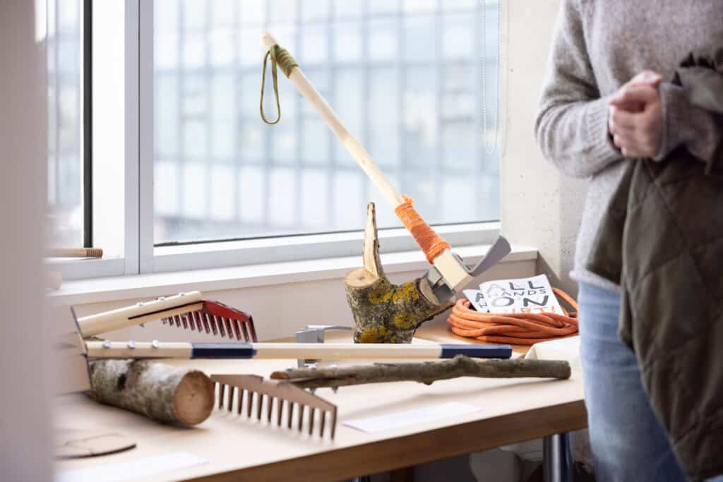 Handmade trail tools rest on a table beside branches and carved wood, bringing the textures of the forest indoors during a community studio display.