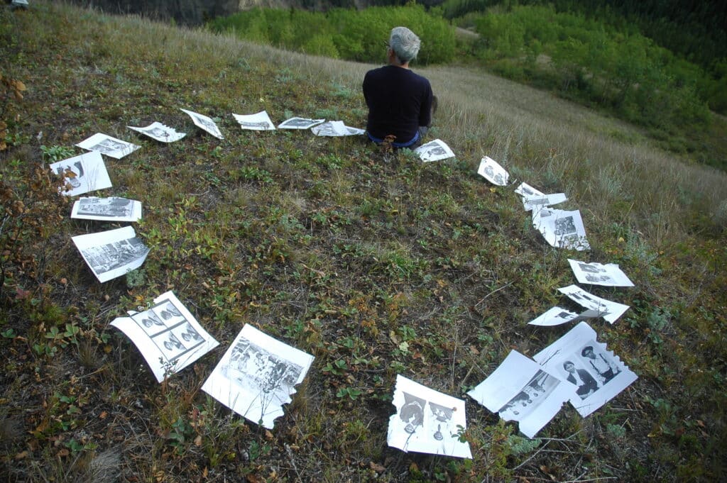 A person sits alone on a grassy hillside encircled by scattered printed photographs, as the landscape opens quietly around them.
