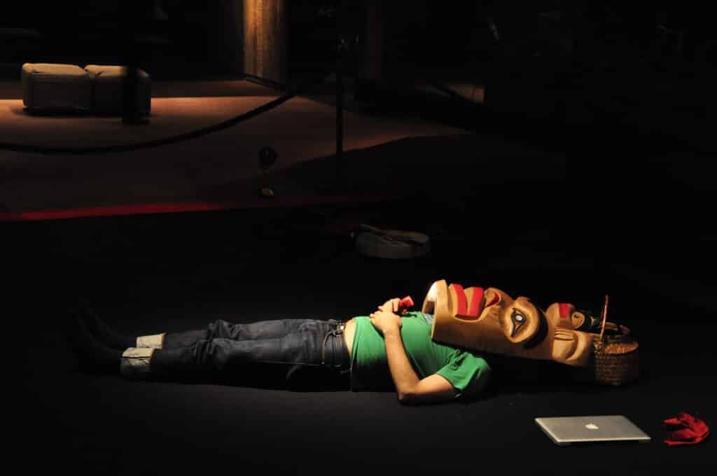 In a dim gallery space, a person lies on the floor beneath a carved mask, illuminated by a single pool of warm light beside everyday objects and technology.