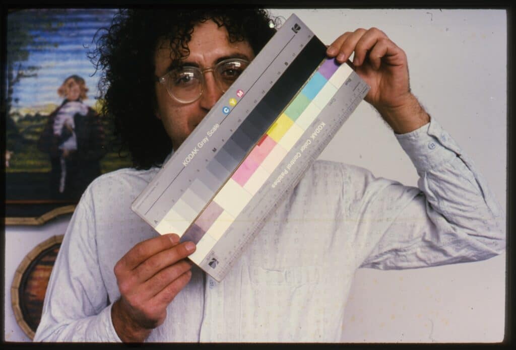 A person with curly hair and glasses holds a Kodak colour calibration chart in front of their face, standing beside framed artwork in a studio or gallery setting.