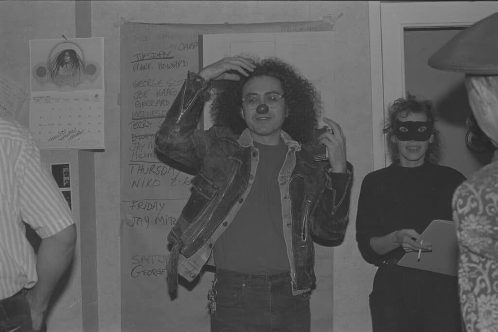 Black-and-white photograph of a person wearing a clown nose and denim jacket gesturing animatedly in a gallery or studio space, while another masked attendee stands nearby holding a clipboard.