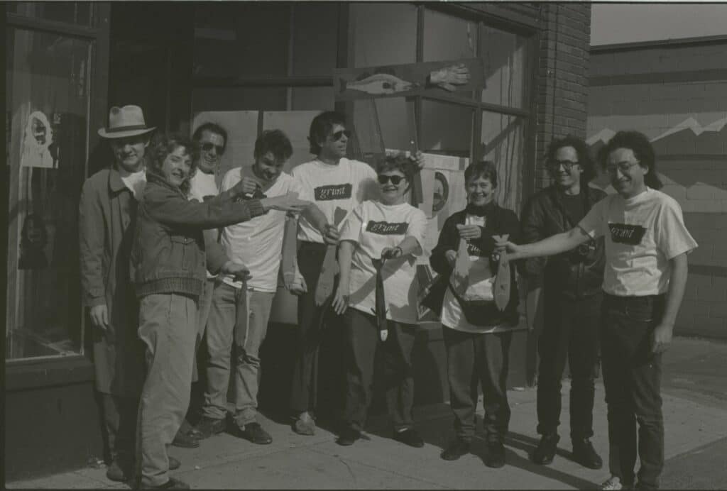 A group of people stand together on a sidewalk outside a gallery storefront, smiling and gesturing toward one another during what appears to be a community art event.