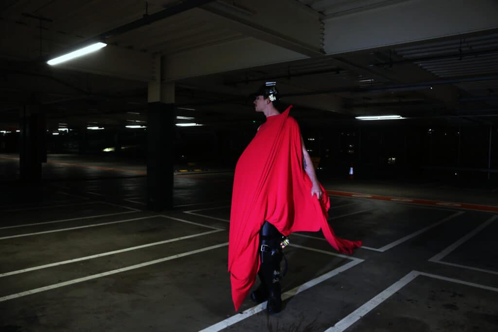 A model walks through a dim parking garage wearing a flowing red draped garment that trails behind them, illuminated by overhead industrial lighting.
