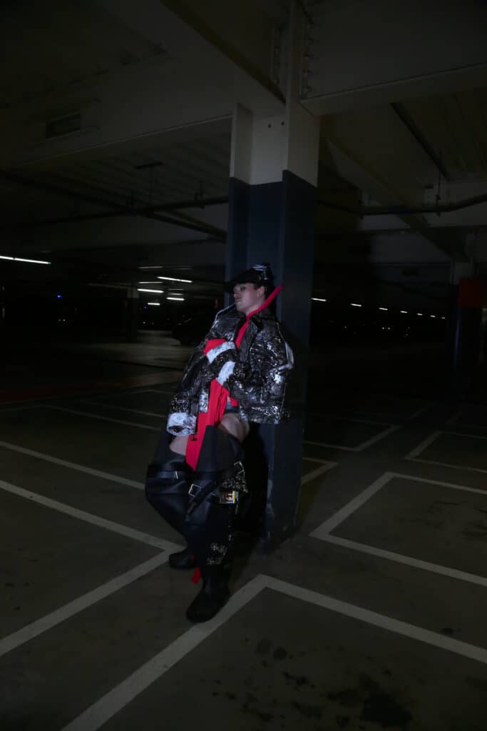 A model leans against a column in a dark parking garage wearing a metallic textured jacket, vivid red draped fabric and oversized black accessories, creating a striking contrast of colour and texture.