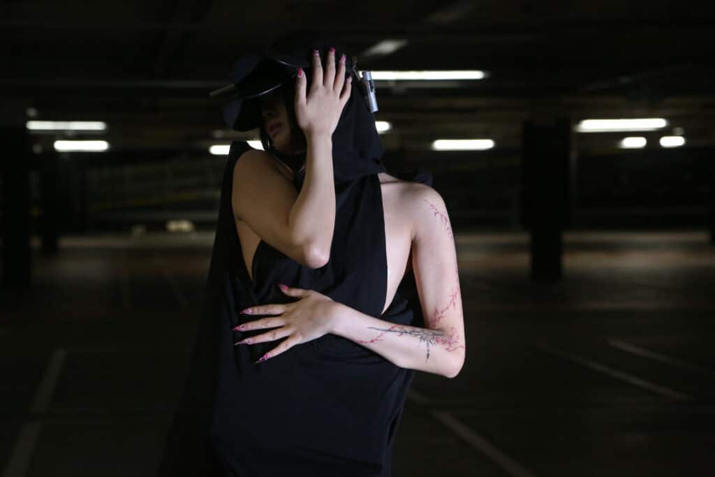 A model in a draped black garment and visor-like headpiece poses in a dimly lit parking garage, with one hand raised to the headpiece and the other resting across the torso.