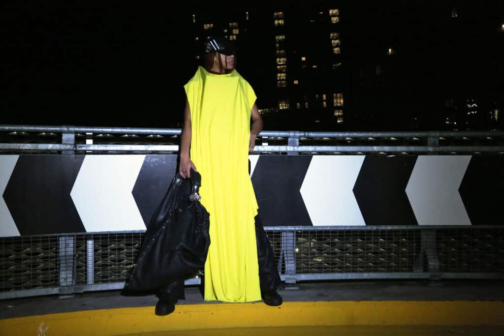 A model wearing a bright neon yellow draped garment and oversized black bag stands on a rooftop parking structure at night, illuminated against a backdrop of city lights.