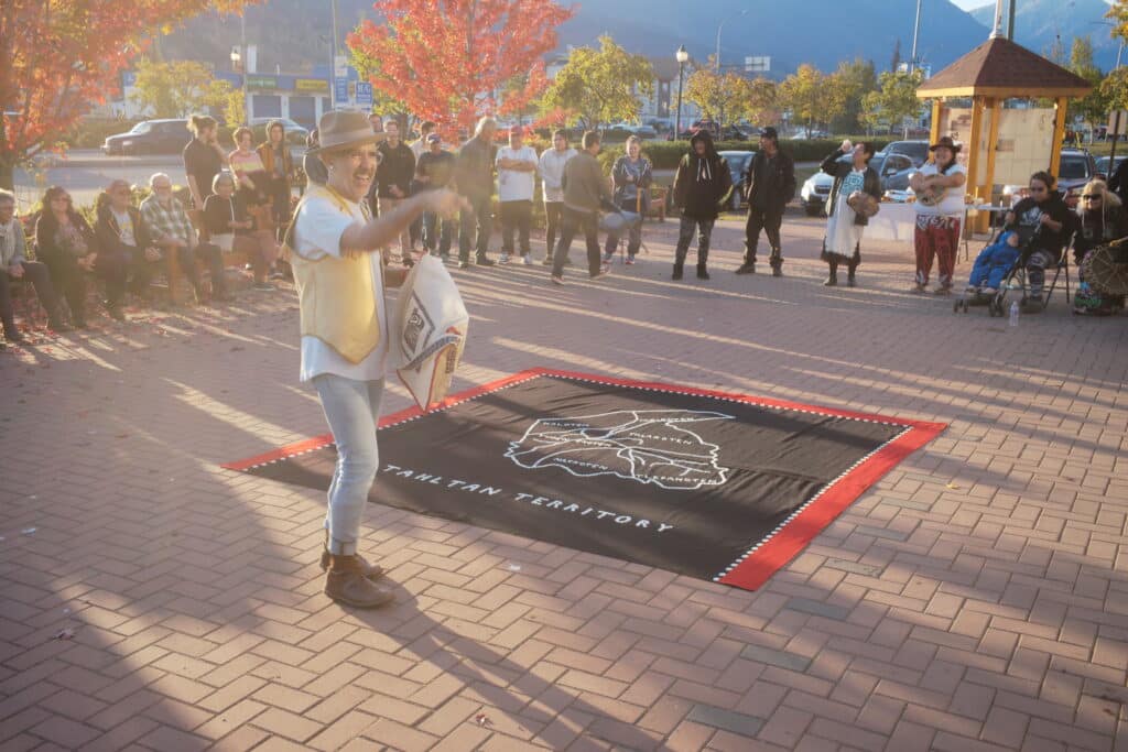 A community gathers in an open plaza during golden evening light, surrounding a speaker beside a large textile marked “Tahltan Territory” as mountains rise in the distance.
