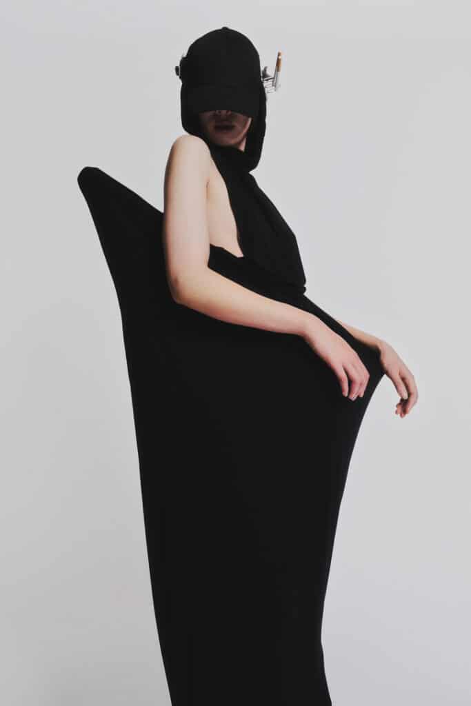 A model wears a flowing black sculptural garment and visor-like headpiece against a light grey background, creating a dramatic silhouette with angular and elongated forms.
