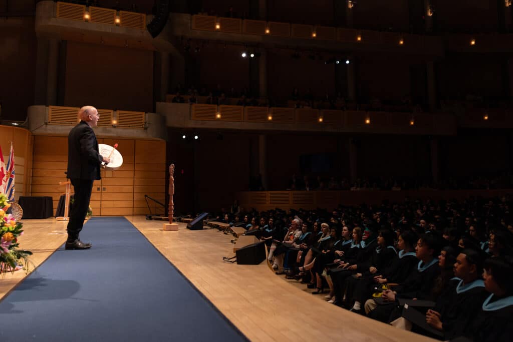 A speaker holding a hand drum stands before graduates in a warmly lit concert hall, as balcony lights glow above the audience like constellations in the dark.