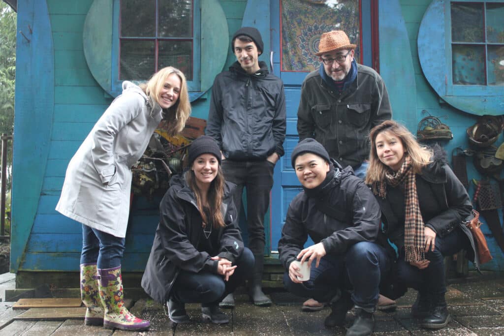 Six people pose in front of a bright blue cabin on a rainy day, bundled in jackets and hats, with painted shutters and eclectic details framing the scene.