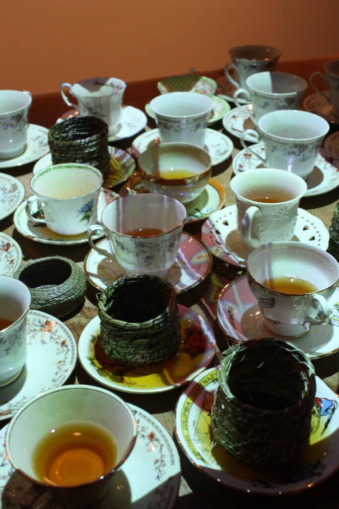A collection of teacups and saucers filled with amber tea gathers across a table, while woven vessels rest among them.