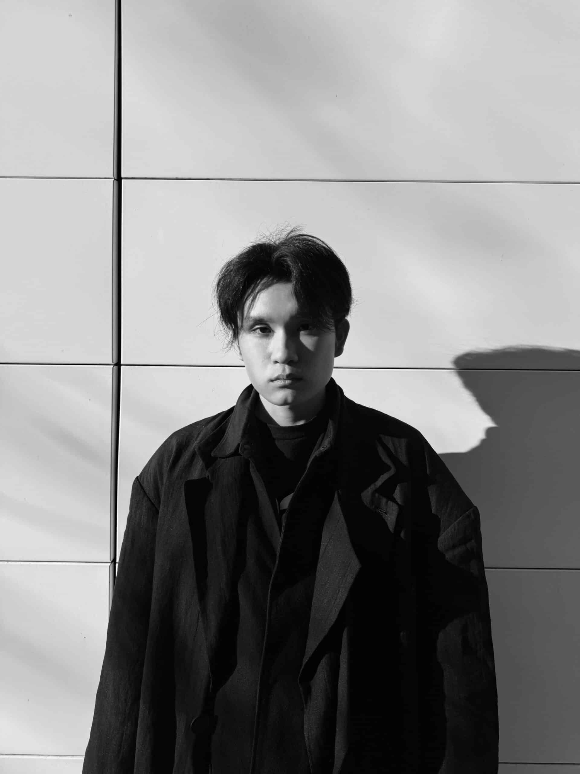 A black and white photo of a person standing behind a light background, in a black coat.