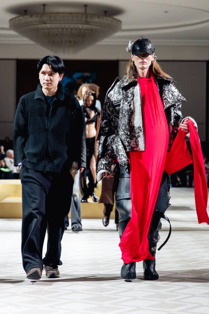 A fashion designer walks beside a runway model wearing a metallic textured jacket and bright red draped garment during a fashion presentation, with other models visible in the background.
