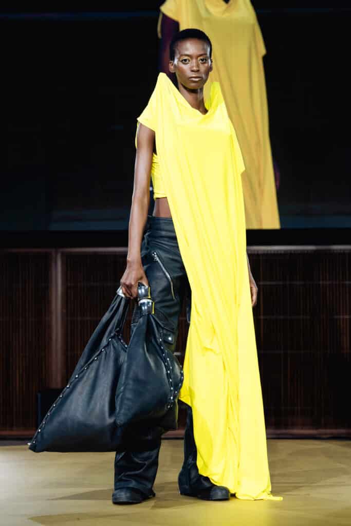 A runway model wears a bright yellow draped garment paired with black leather trousers and carries an oversized black bag while walking down the catwalk.