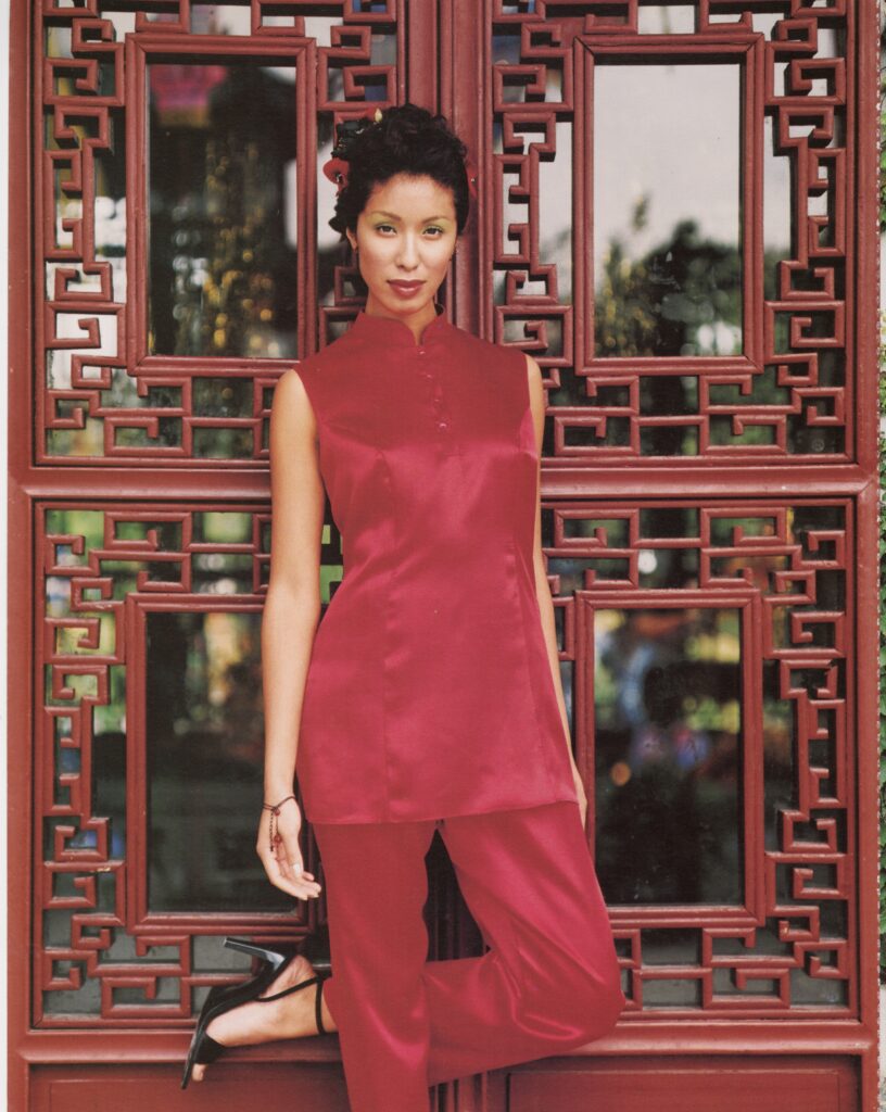 A model poses in a red silk tunic and trousers in front of an ornate lattice screen, blending fashion with architectural detail.