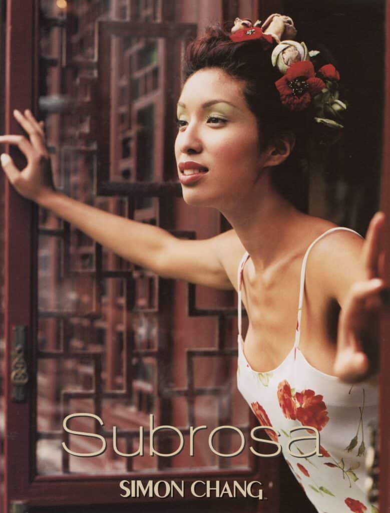 A model wearing a floral dress and flower-adorned hairstyle leans through a decorative doorway in a fashion campaign photograph.