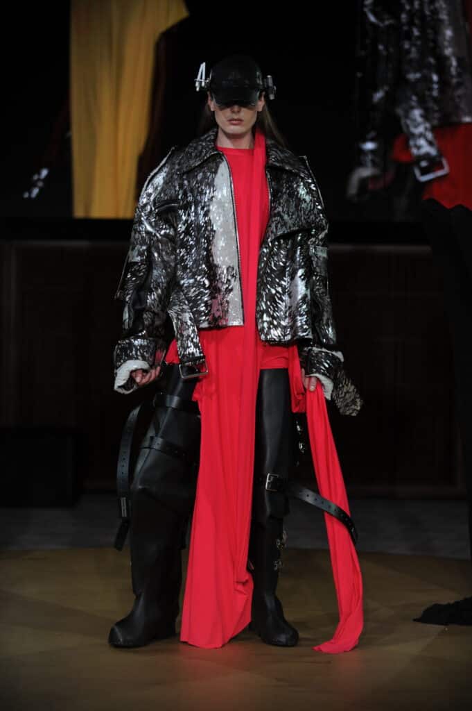 A model walks the runway wearing a metallic black-and-silver jacket layered over vivid red draped fabric, paired with oversized black accessories and a futuristic headpiece.