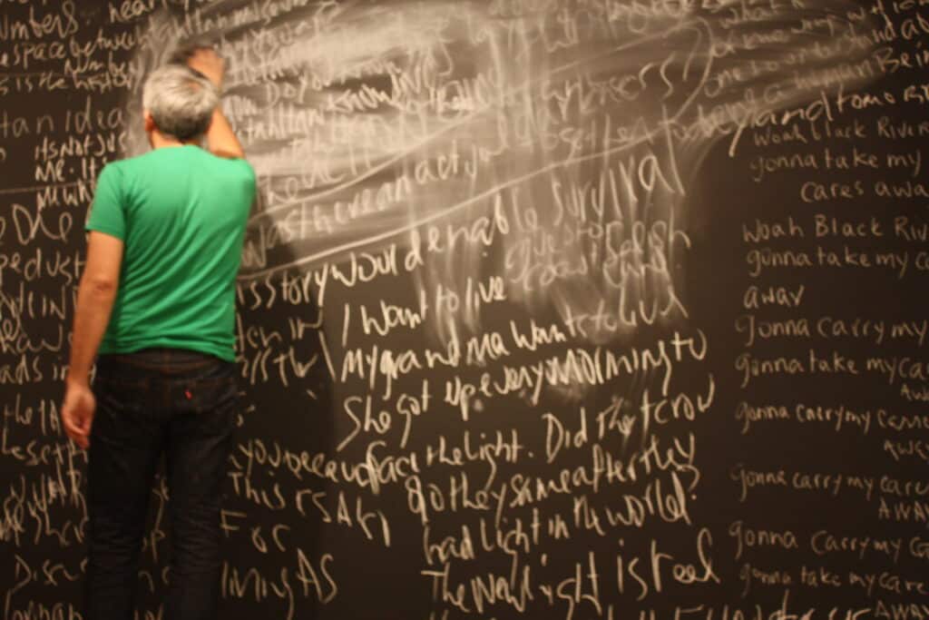 A person writes across a wall-sized chalkboard covered in layered words and sketches.