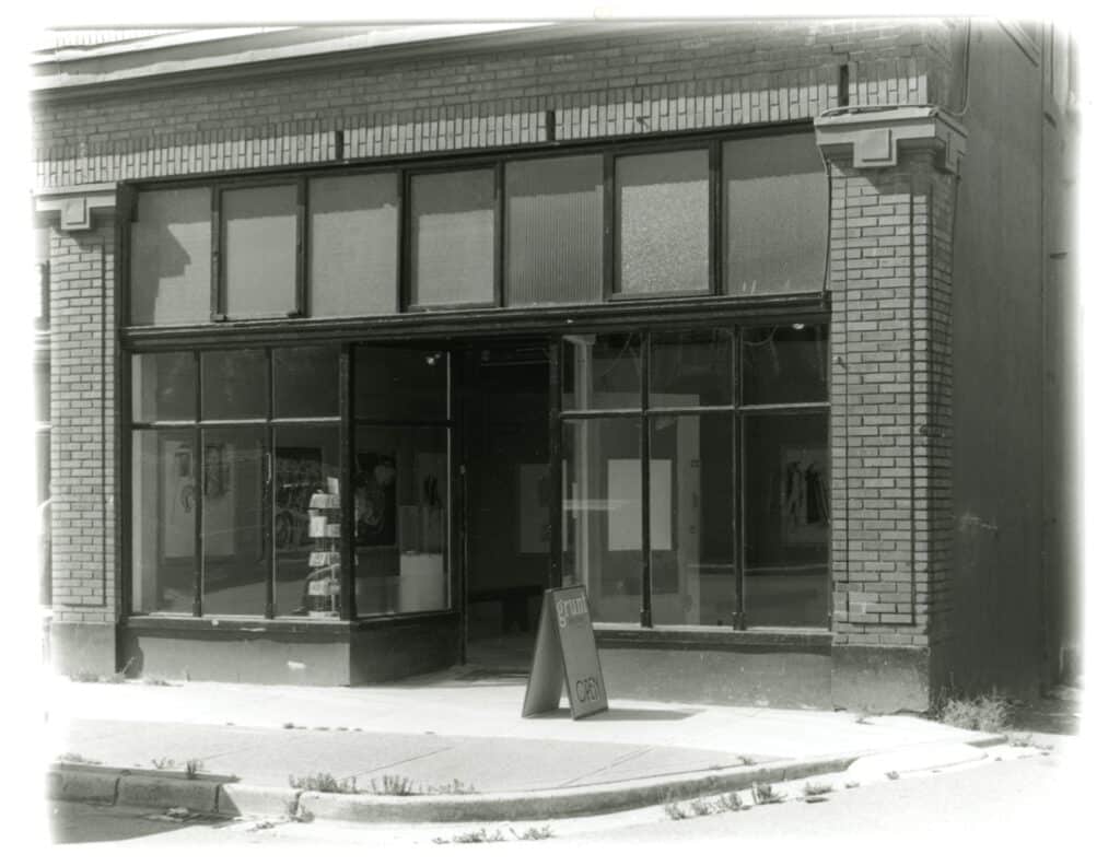 Black-and-white photograph of a corner gallery space with large industrial windows and an open entrance, viewed from across a quiet street.