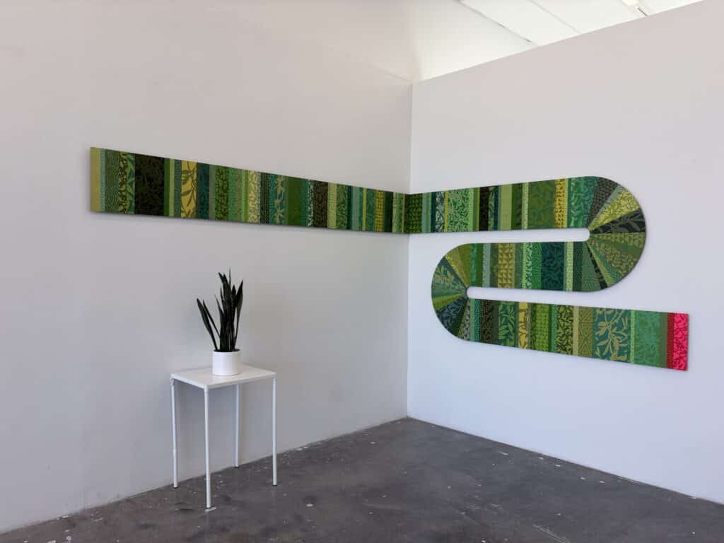 A long green artwork wraps around the corner of a white gallery wall, weaving together layered botanical patterns and geometric motifs. A small potted plant on a white table echoes the work's organic palette.